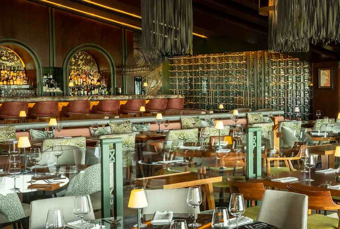 Interior view of a spacious, opulent restaurant dining room in Cancún set for large-scale service, featuring a prominent, fully-stocked bar with arched details, a backlit wine display, and unique, high-end chandelier fixtures. The layout accommodates numerous tables with elegant place settings, making it an ideal venue for MICE industry gala dinners, large corporate banquets, and sophisticated incentive travel group dining